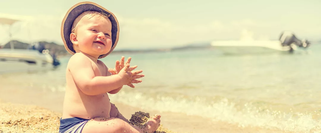 Bébé qui rigole sur le sable assis devant la mer avec un chapeau